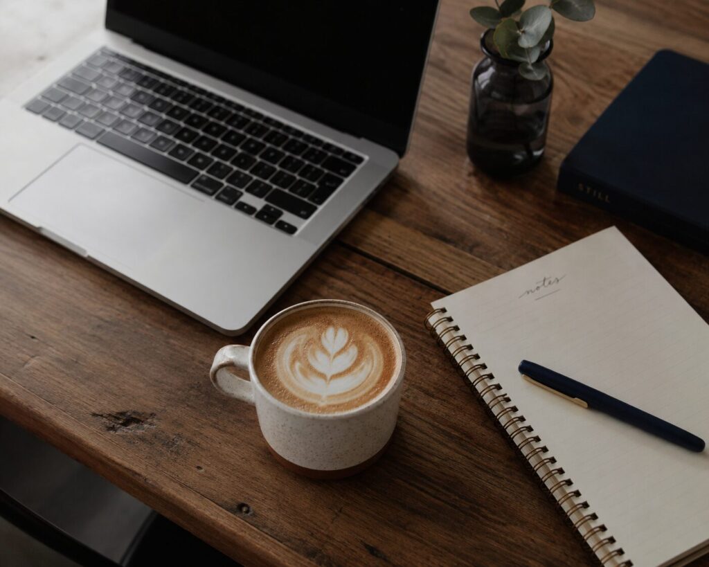 Angled view of a wooden table with an open laptop, latte, spiral notebook, and navy pen, creating a warm, minimalist workspace with subtle navy accents.